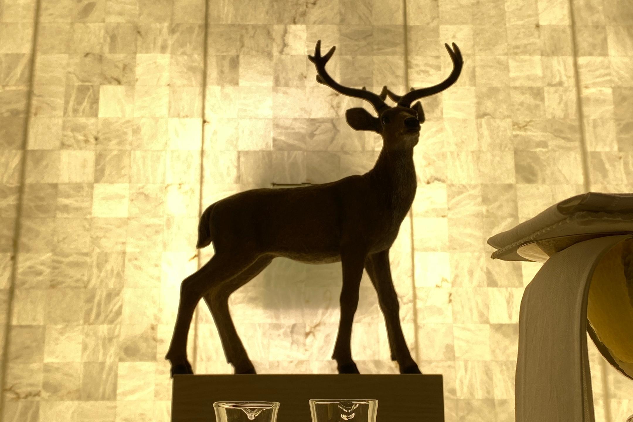 Hirschfigur auf Sockel, davor ein Altar mit Wasser und Wein, Hostienschale und Kelch. Die Wand im Hintergrund erstrahlt in goldenem Licht.